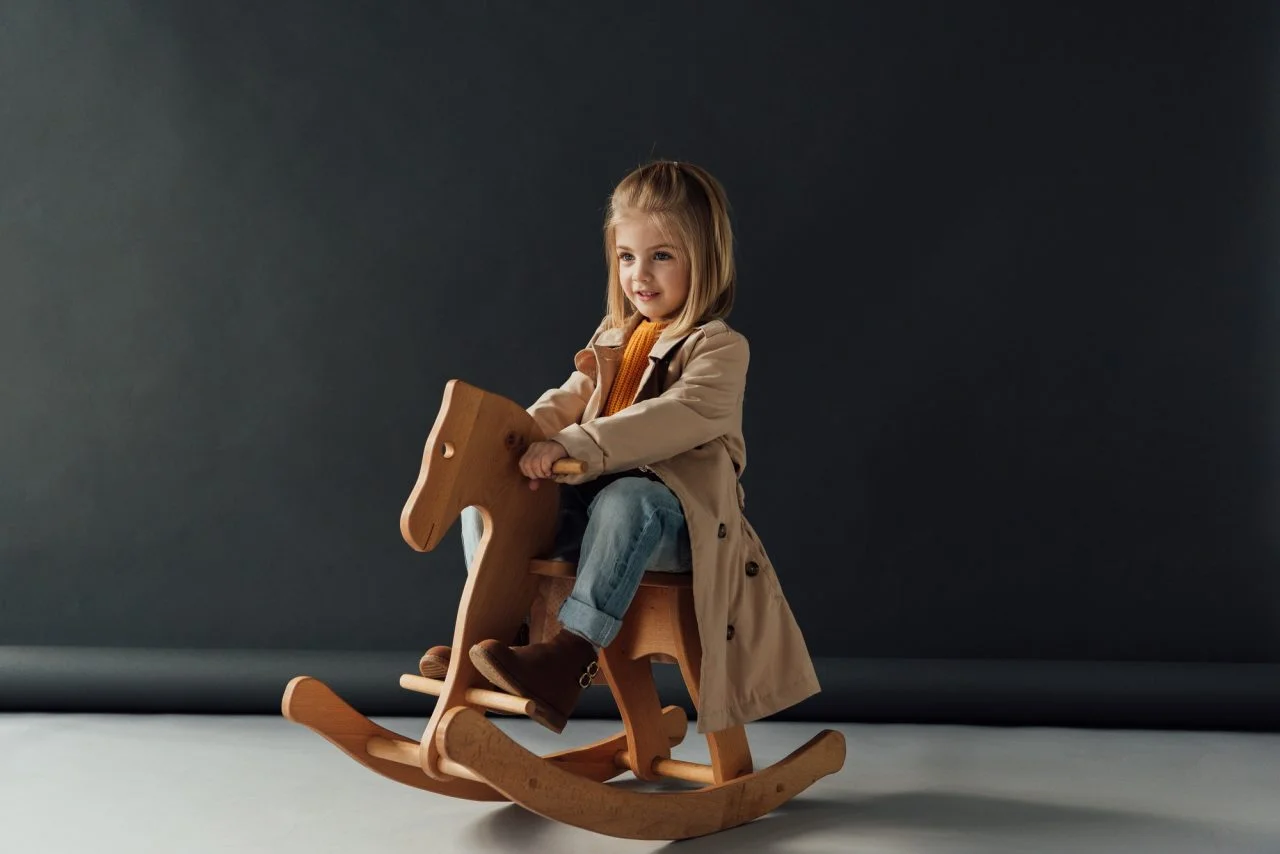 smiling-child-in-trench-coat-and-jeans-sitting-on-rocking-horse-on-black-background